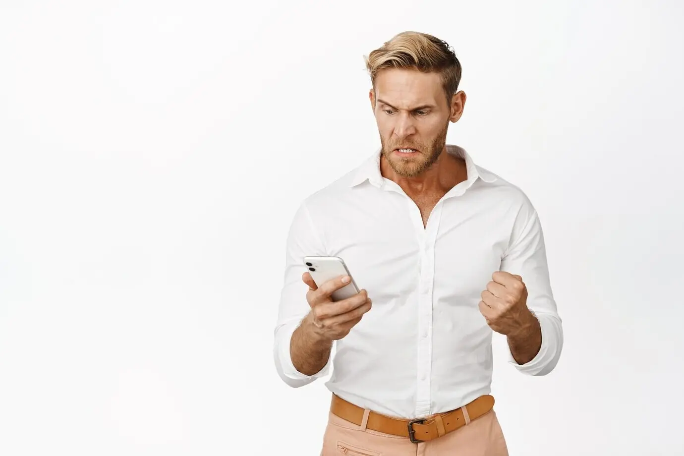 Un homme en colère regarde son téléphone portable, avec une expression du visage exaspérée et furieuse, frustré par quelque chose sur son téléphone, debout sur fond blanc.